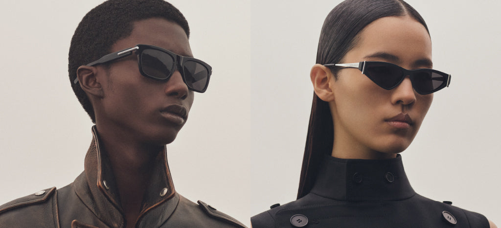 Two models wearing black sunglasses and high-collared, dark-colored jackets. The left model has short hair, while the right model has long, straight hair pulled back. Both have neutral expressions against a plain background.