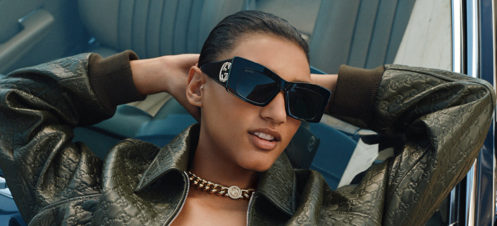 A person wearing large black sunglasses, a gold chain necklace, and a green textured jacket leans back in a car with hands behind their head, exuding a confident, stylish vibe.