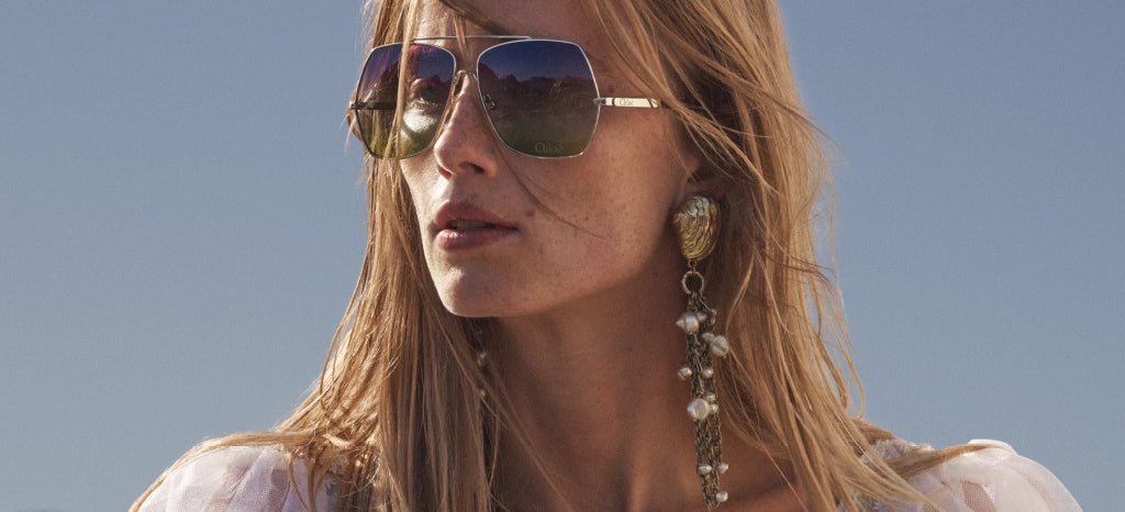 A woman with long blonde hair wears large square sunglasses and a long, dangling earring decorated with pearls and gold. She is outdoors under a clear blue sky, looking slightly to the left.