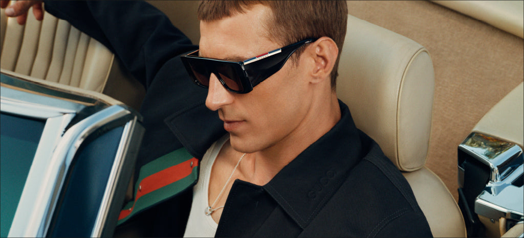 A man wearing large, stylish black sunglasses and a dark jacket sits in the drivers seat of a beige convertible car, looking down slightly with a relaxed expression.