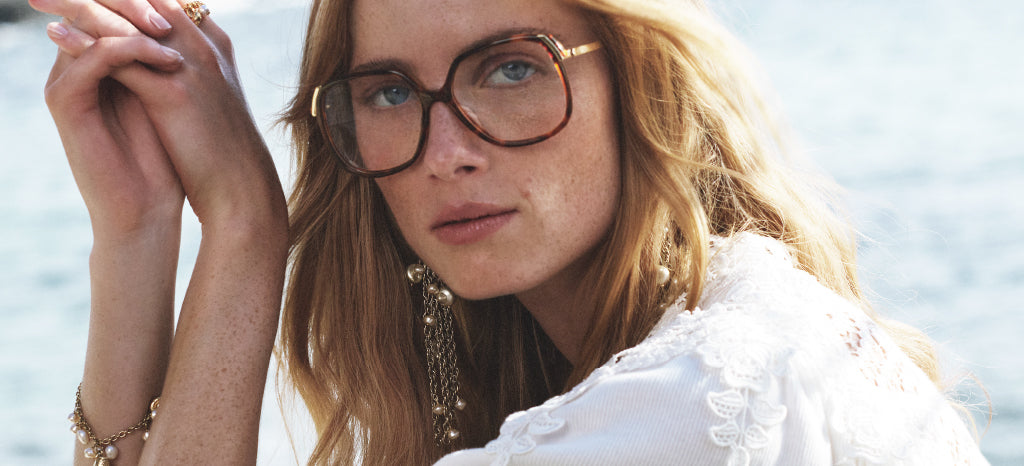 A woman with long blond hair wears oversized brown glasses, a white lace top, and gold jewelry, posing outdoors with a calm body of water in the background.