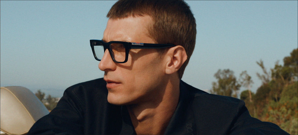 A man with short light brown hair wearing black rectangular glasses and a dark jacket sits in a car outdoors, looking to the left with trees and a clear sky in the background.