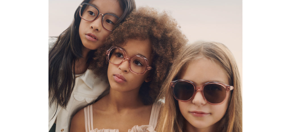 Three girls wearing stylish eyeglasses and sunglasses pose closely together, looking thoughtfully in different directions against a soft, neutral background.
