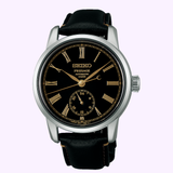 A Seiko Luxe Watches Presage Craftsmanship Series Enamel SPB499 automatic wristwatch featuring an elegant black enamel dial, gold Roman numerals, a subdial at 6 o’clock, and a refined black leather strap. The case is silver with a ridged crown on the right side—an exquisite example of Seiko Luxe Watches’ renowned craftsmanship, powered by an automatic movement.