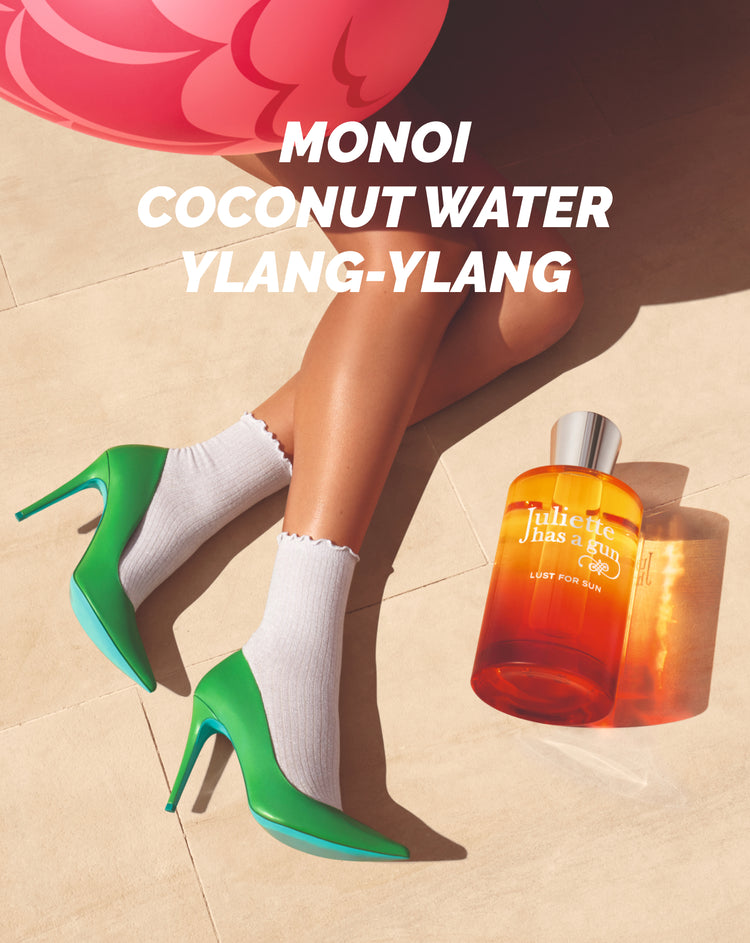 A person wearing green high heels and white socks sits with legs crossed beside an orange perfume bottle labeled Juliette Has a Gun. Bold text above reads: MONOI COCONUT WATER YLANG-YLANG.