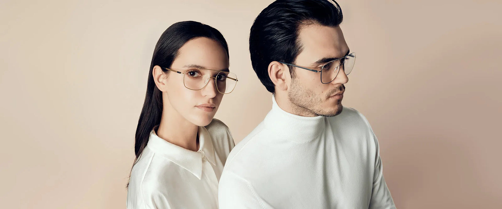 A woman and a man wearing stylish eyeglasses and white clothing stand back to back against a light beige background, gazing confidently toward the camera.
