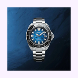 The Seiko Watches SRPE33 Prospex Men watch features a silver stainless steel strap, blue analog dial, black bezel, and date display at 3 o'clock—all shown against a textured blue background.