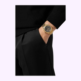 A person dressed in black clothing has their hand in their pocket, displaying a Versace Watches VE6A00623 V-Code men’s watch with a gold stainless steel band and a sleek black face.
