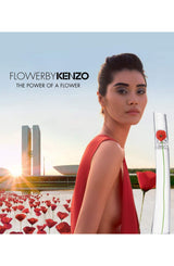 A woman in a red dress stands among poppies by water at sunset. In front, a Flower By Kenzo 3.4 oz EDP for Woman Perfume bottle by Kenzo Perfumes displays a red flower design. Text: Flower By Kenzo—The Power of a Flower.