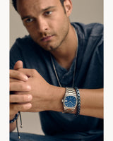 A man in a blue shirt sits with his hands clasped, wearing a Bulova Watches 96B440 Super Seville Men (UPC 042429599789) and a black beaded bracelet. The softly lit, neutral background highlights the elegance of Bulova Watches.