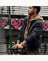 A man in a brown hoodie and dark jacket stands by a bicycle with graffiti art behind him, reflecting the bold urban style of Bulova Watches 96B440 Super Seville Men (UPC 042429599789).