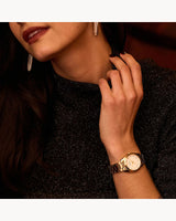 A woman with long dark hair and dangling earrings touches her neck, wearing a sparkly black top and a Bulova Watches 97P172 Gold Stainless Steel Strap Lady Watch with a white face on her left wrist. Only her lower face and hand are visible.