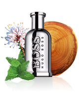 A silver Hugo Boss Perfumes Bottled United 3.3 oz EDT for Men is pictured with a wood disc, mint, and flowering herbs, highlighting its natural fragrance notes.