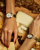 Two hands, one with red nail polish, rest on a wooden board with cheese, crackers, grapes, and dates. Both wear Bulova 97P172 Gold Stainless Steel Strap Lady Watches featuring white faces and metal bands by Bulova Watches.