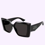 Black, oversized square sunglasses with thick frames and dark lenses. The Alexander McQueen Sunglasses AM0467S-001 56 feature the brand name printed in white on one of the arms, showcasing the designer’s signature eyewear style. Set against a light pastel background.