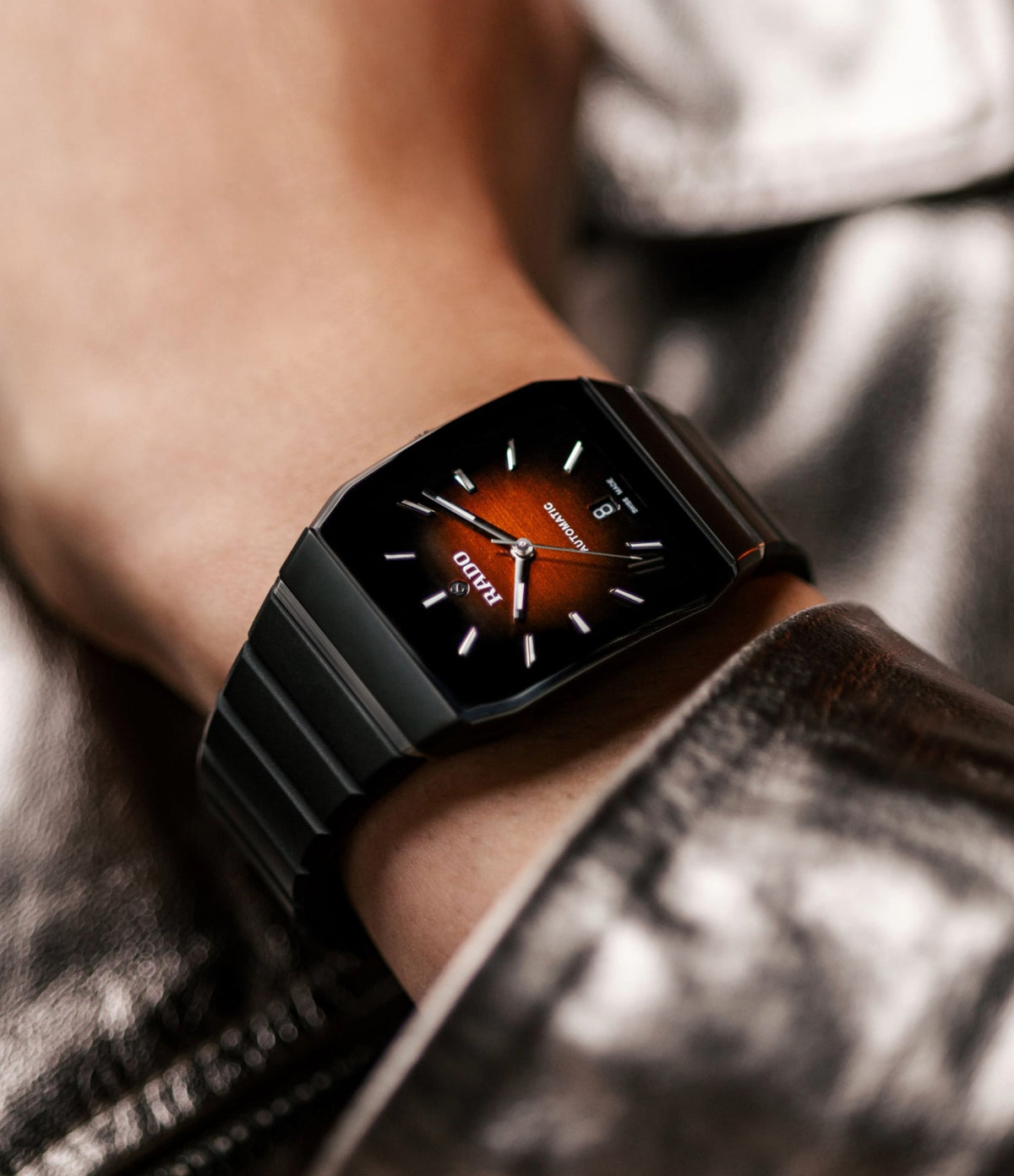 A close-up of a person wearing a black RADO watch with a red gradient dial and white hour markers, dressed in a shiny metallic silver jacket.
