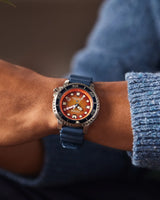 A person in a blue sweater showcases the Citizen BN0169-03X Eco-Drive Men's Watch by Citizen Watches, featuring a stainless steel case, blue strap, vivid orange dial with bold white markers, and a silver bezel.