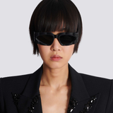 A person with short, straight black hair wears Balmain Sunglasses B-Spirit BPS-179A-56 Unisex and a black sequin blazer, posing against a plain light gray background.