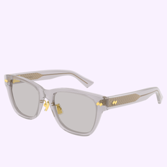 Bottega Veneta Sunglasses BV1402SK-004 55 feature clear frames, rectangular grey lenses, gold metal accents on the hinges and temples, and a textured gold pattern on the sides, shown against a light purple background.