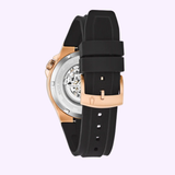 The Bulova 98A177 Maquina Automatic by Bulova Watches features a black silicone strap, rose gold buckle, and an exhibition caseback revealing the mechanical movement against a light pastel background.
