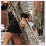 A person in a long checkered coat and black textured skirt, wearing the Bulova Watches Goddess of Time Sophia 98P237 gold-silver watch, crosses a city street with blurred buildings and cars in the background; their face is not visible.