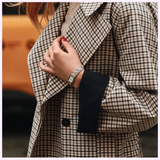 A person in a plaid coat with black trim displays red nail polish, a silver bracelet, and a Bulova Watches Goddess of Time Sophia 98P237, with a blurred yellow background adding to the stylish vibe.