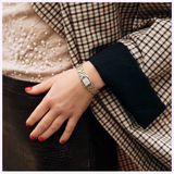 A person with red nail polish in a pearl top, checkered blazer, and black skirt rests their hand on their hip, showcasing a Bulova Watches Goddess of Time Sophia 98P237 gold and silver women's watch.