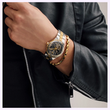 A person in a black leather jacket displays the Bulova Watches Prestige Automatic 98A339, featuring a black dial, gold and silver-tone stainless steel, and an automatic movement, paired with a gold chain bracelet near the zipper.