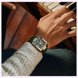 A person wearing a Bulova Watches Series X Automatic 98A343 with a green square face plays piano keys, the luxury men's watch standing out against their light knit sweater and blue pants.