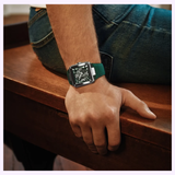 A person in blue jeans sits on wood, hand resting nearby, wearing a Bulova Watches Series X Automatic 98A343 with a green strap and striking square dial that reveals its skeletonized automatic movement.