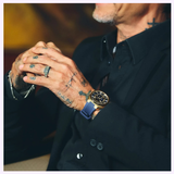 A person in a dark suit with facial hair and tattooed hands sits with interlaced fingers, wearing a ring and the Bulova Watch Surveyor Marc Anthony 97A205, featuring automatic movement, blue strap, and gold-tone stainless steel details.