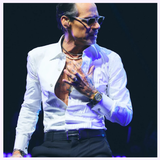 A man in sunglasses, an open white dress shirt, black pants, and jewelry—including a Bulova Watches Surveyor Marc Anthony 97A205—stands on stage with one hand on his chest, looking to the side under blue lighting.