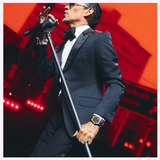 A man in a tuxedo sings passionately on stage, wearing the Bulova Watch Surveyor Marc Anthony 97A205 by Bulova Watches, as dramatic lighting and a vivid red backdrop amplify his performance.