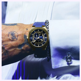 A close-up of a tattooed wrist wearing the Bulova Watches Surveyor Marc Anthony 97A205, featuring gold-tone stainless steel and navy blue accents, the hand dressed in a white shirt with a cufflink.
