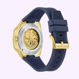 The Bulova Watches Surveyor Marc Anthony 97A205 features a gold-tone stainless steel case, navy blue rubber strap, and a transparent case back that reveals the automatic movement and mechanical details.