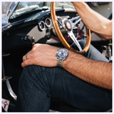 A man wearing a Bulova Watches Shelby Daytona coupe 96K118 Limited Edition watch and jeans sits in the driver’s seat of a classic car, holding the wooden steering wheel with one hand.