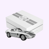 A small silver model car is positioned in front of a white Bulova Watches gift box with gray stripes and the Shelby logo on the lid, celebrating the Bulova Watches Shelby Daytona Coupe 96K118 Limited Edition.