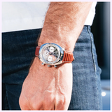 A close-up of a person’s wrist wearing a Bulova Watches Shelby Daytona Coupe 96K118 Limited Edition, a silver chronograph timepiece with a brown leather strap, set against blue jeans and a white shirt.