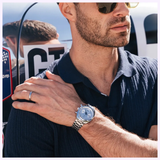 A man wearing sunglasses and a blue polo shirt stands by a car adorned with racing stickers on the window, showcasing his Bulova Watches Shelby Daytona Coupe 96K118 Limited Edition watch and a wedding ring on his left hand.