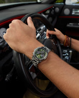 Wearing the Citizen Watches CA4664-60W Promaster Navihawk, a person with brown beaded bracelets grips a black steering wheel in a car with black and red interior accents.
