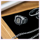 A silver CASIO RING WATCH CRW001-1 from G-Shock Watches is displayed on a black surface alongside metal rings and a chain, with the corner of a closed book visible in the background, creating an elegant arrangement that highlights the timeless appeal of this distinctive timepiece.