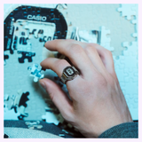 A hand wearing the G-Shock Watches CASIO RING WATCH CRW001-1—a distinctive rectangular digital ring watch—rests on an unfinished jigsaw puzzle, with loose pieces and a partially visible Casio logo in the background.