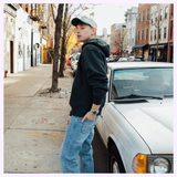 A man in a black hoodie, jeans, and a cap stands beside a white vintage car on a city street, glancing back over his shoulder. On his wrist, the G-Shock Watches CASIO G-Shock Watch Analog-Digital GA010-5A with Carbon Core Guard construction and 10-year battery life adds a touch of modern durability to his classic style.