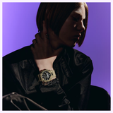 A person with chin-length hair, dressed in a black shirt, sits against a purple background wearing the G-Shock Watches CASIO G-Shock Watch Analog-Digital GA010-5A featuring Carbon Core Guard technology, while touching their neck with one hand.