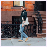 A young man in jeans, a black hoodie, and tan shoes walks along a city sidewalk, carrying a beige tote bag and wearing the CASIO G-Shock Watch Analog-Digital GA010-5A from G-Shock Watches. Brick buildings, stairs, and a bicycle appear in the background.