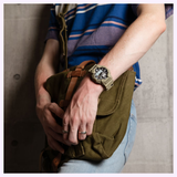 A person wearing a striped shirt and jeans holds an olive green bag. On their left hand, they wear rings and the G-Shock Watches CASIO G-Shock Watch Analog-Digital GA010-5A, featuring Carbon Core Guard technology and a 10-year battery life. The background is a concrete wall.