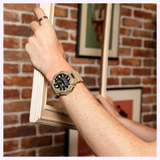 A person wearing a rugged beige CASIO G-Shock Watch Analog-Digital GA010-5A featuring Carbon Core Guard technology by G-Shock Watches holds a white picture frame against a brick wall adorned with framed art.