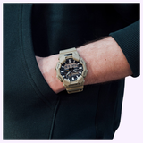 A person wearing a green sweatshirt has their hand in their pocket, showing off a beige CASIO G-Shock Watch Analog-Digital GA010-5A by G-Shock Watches on their wrist, which features both analog and digital displays along with Carbon Core Guard construction for enhanced durability.