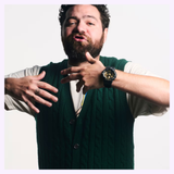 A bearded man in a green sweater vest, wearing a cream-colored shirt and a CASIO G-Shock Watch Bold Camo Series GA700CF-1A from G-Shock Watches, gestures with his hands while looking at the camera, standing against a plain white background.