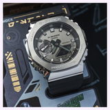 A CASIO G-Shock Watch G-STEEL GM-2100-1ASW with a silver face and black band sits on a sci-fi themed box, featuring bold graphics along with yellow and green text.